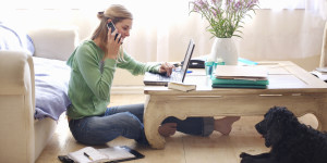 Mid adult woman sitting on floor, leaning on sofa, talking on, using laptop, side view